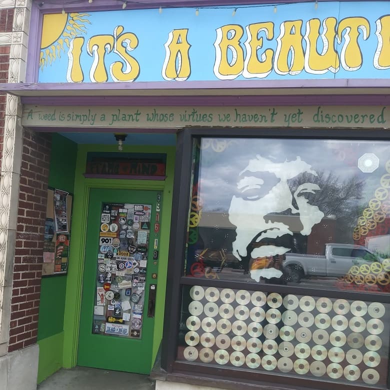 The storefront: a hand-painted yellow and blue sign, a green door covered in stickers, and compact discs filling the lower window.